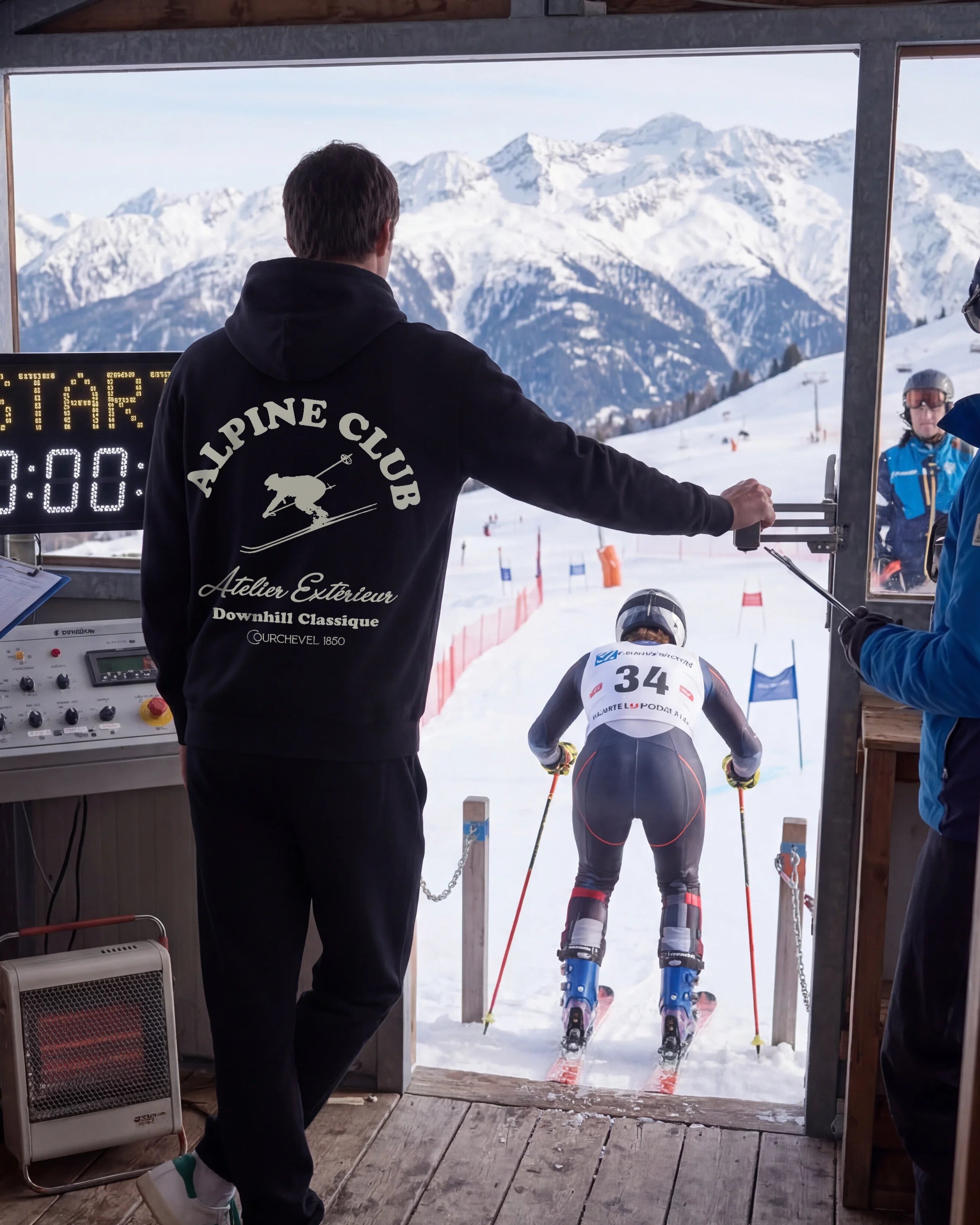 Ski race starting gate with a skier about to begin, watched by officials in a wooden cabin.