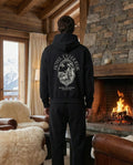 Person wearing a black hoodie with a mountain design in a cozy living room with a fireplace and mountain view.