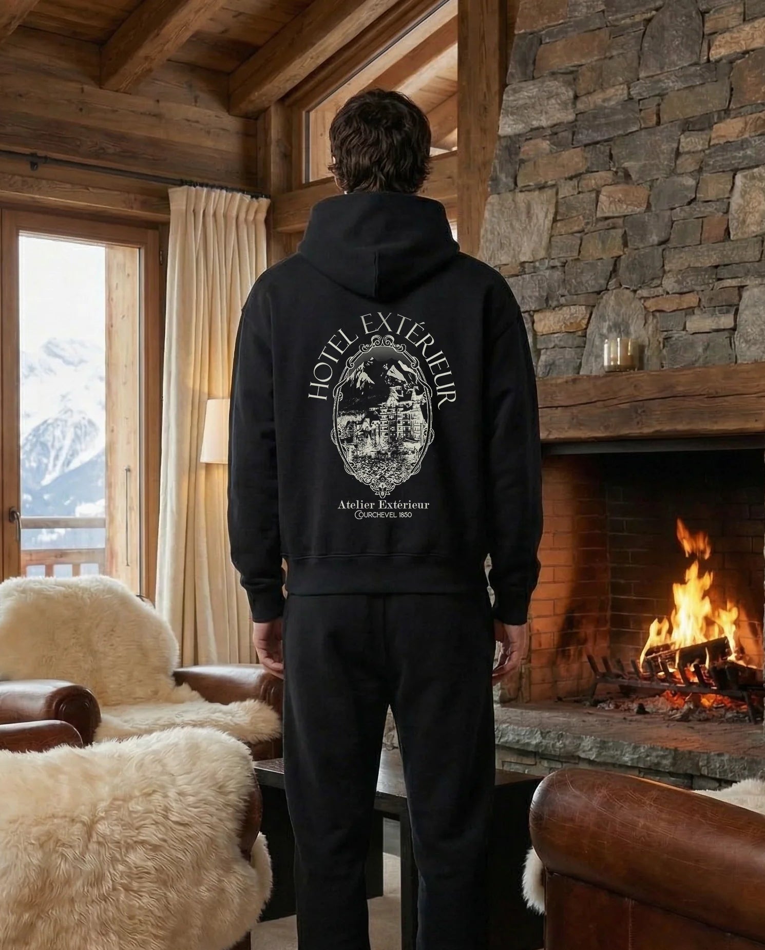 Person wearing a black hoodie with a mountain design in a cozy living room with a fireplace and mountain view.