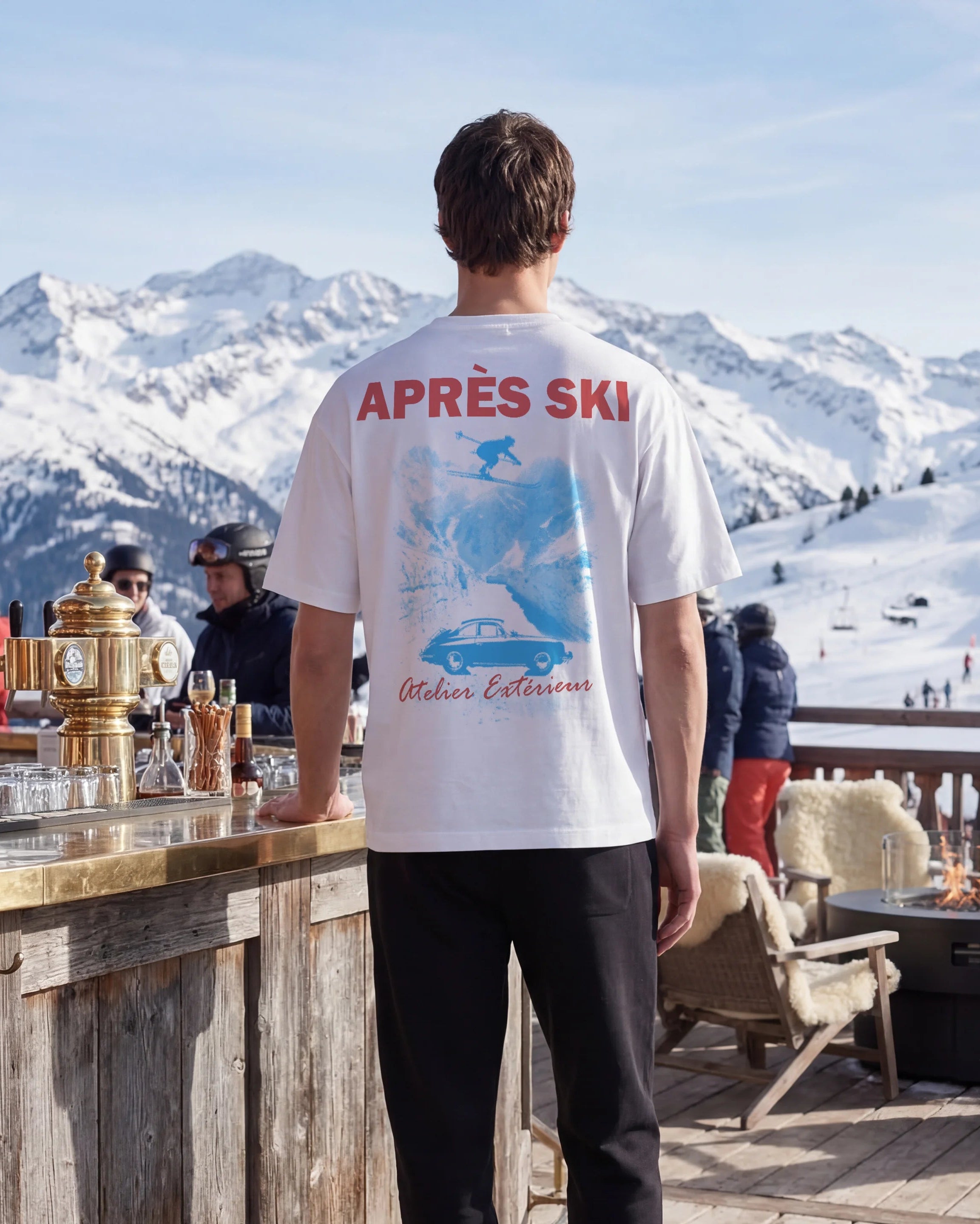 Person wearing a white t-shirt with 'Apres Ski' text and graphic, standing in a snowy mountain resort.