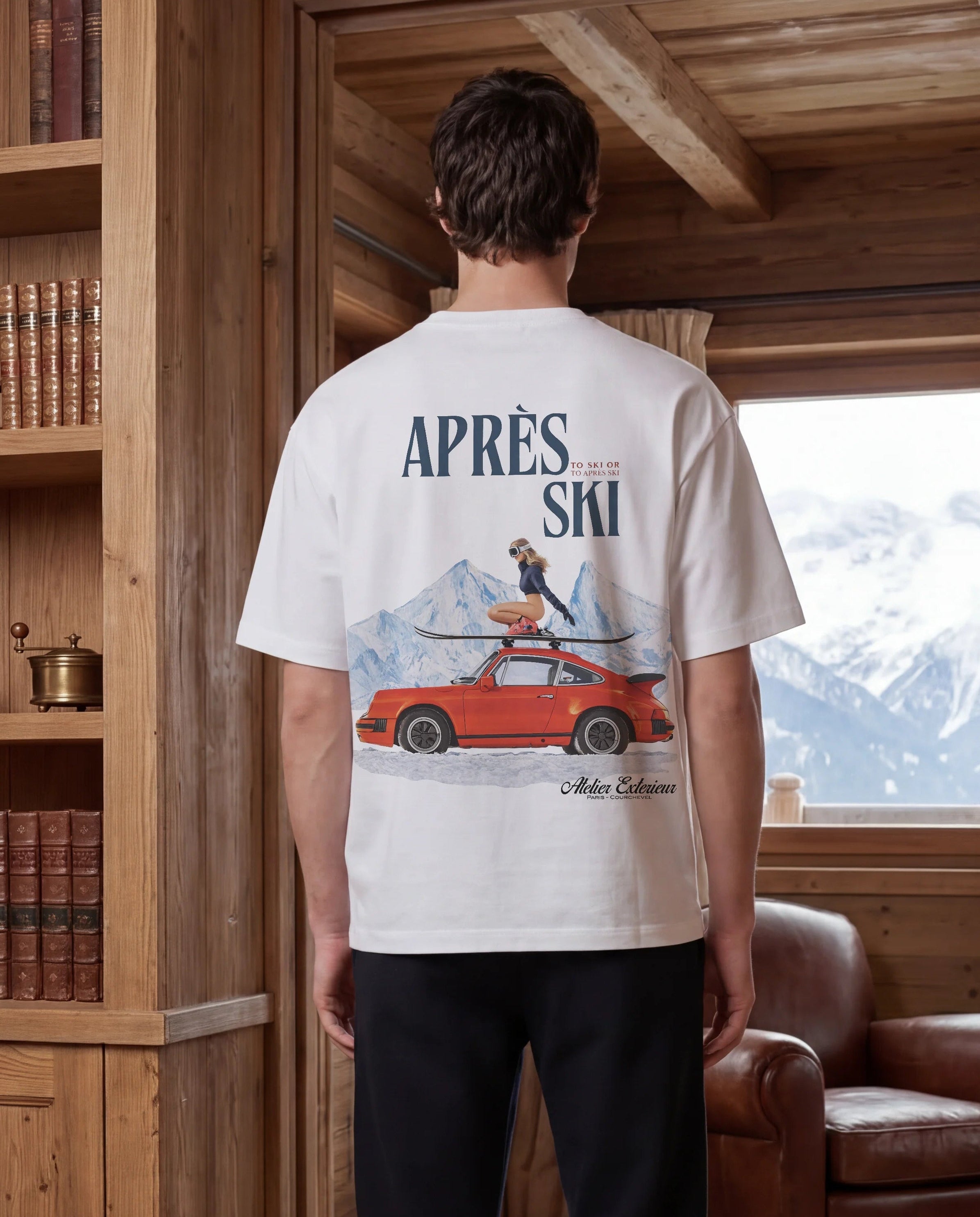 Person wearing a white t-shirt with a graphic design standing in a room with wooden bookshelves and a window view of mountains.