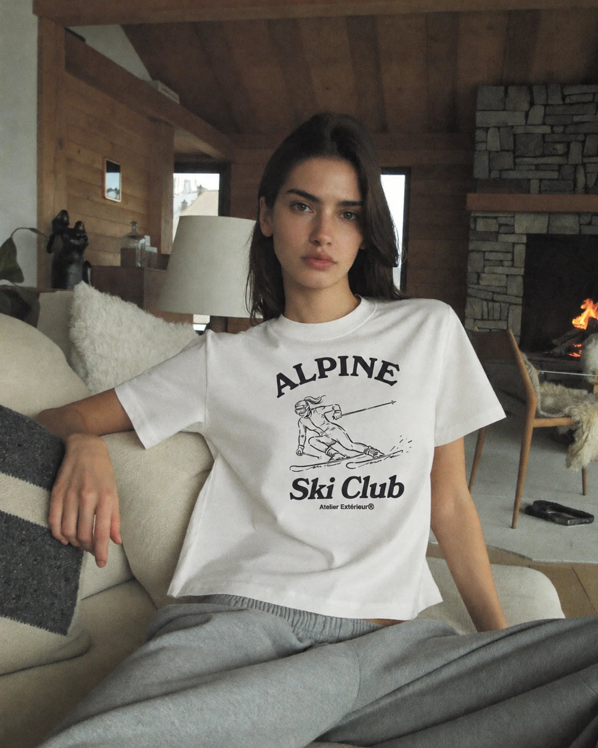 Woman wearing the white Alpine Ski Club tee with grey joggers in a chalet by the fire