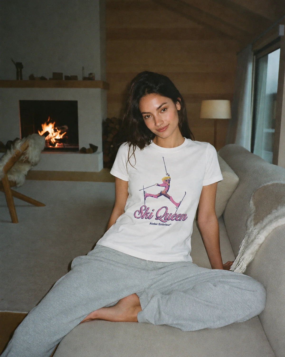 Woman wearing the white Ski Queen tee with grey joggers in a chalet by the fire