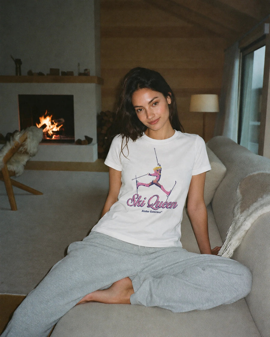 Woman wearing the white Ski Queen tee with grey joggers in a chalet by the fire